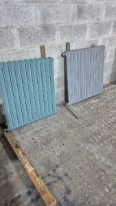 Two ribbed radiators, teal and light gray, propped against a concrete wall in a workshop.