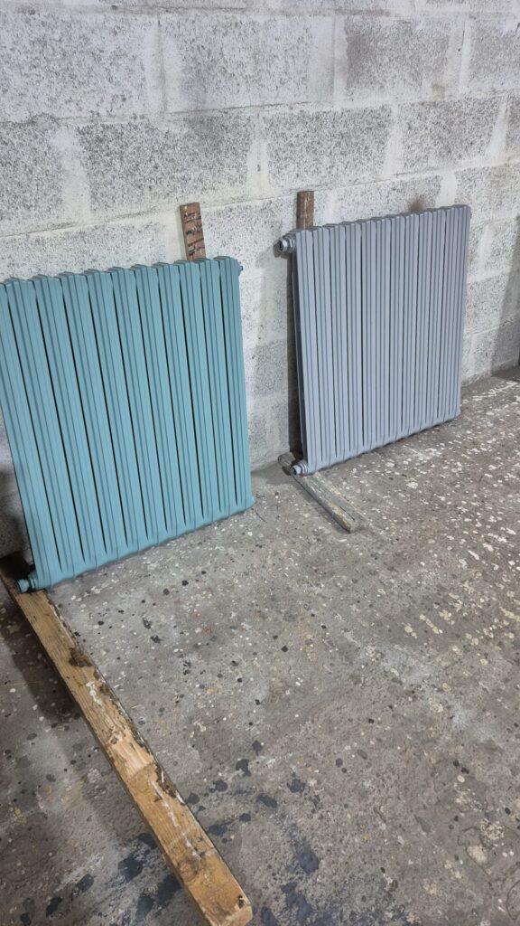 Two ribbed radiators, teal and light gray, propped against a concrete wall in a workshop.